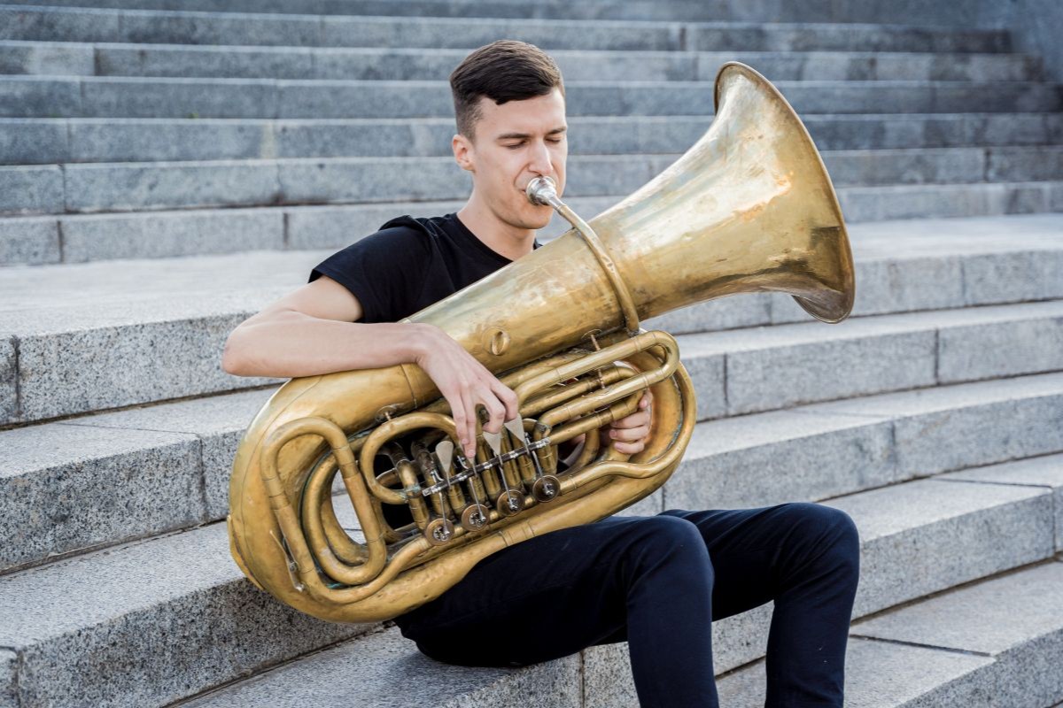 A male playing the Tuba outside sitting on some stairs